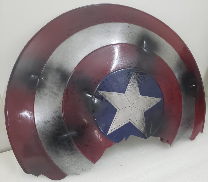 May include: A red, white, and blue Captain America shield with a star in the center. The shield is cracked and damaged, with black paint splatters on the surface.