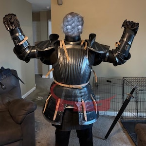 May include: A full suit of black and silver medieval armor with articulated gauntlets and a breastplate. The armor is detailed with leather straps and rivets. A black sword rests on the floor.