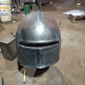 May include: A handcrafted metal helmet with a dark, metallic finish. The helmet features a rounded top with a visor opening and is likely for historical reenactment or display. The helmet is sitting on a metal stand.