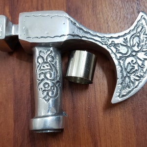 Puede incluir: Una cabeza de hacha de metal de color plateado con un dise&ntilde;o ornamentado. La cabeza del hacha est&aacute; unida a un mango de metal plateado con un dise&ntilde;o decorativo. La cabeza del hacha est&aacute; sobre una superficie de madera.