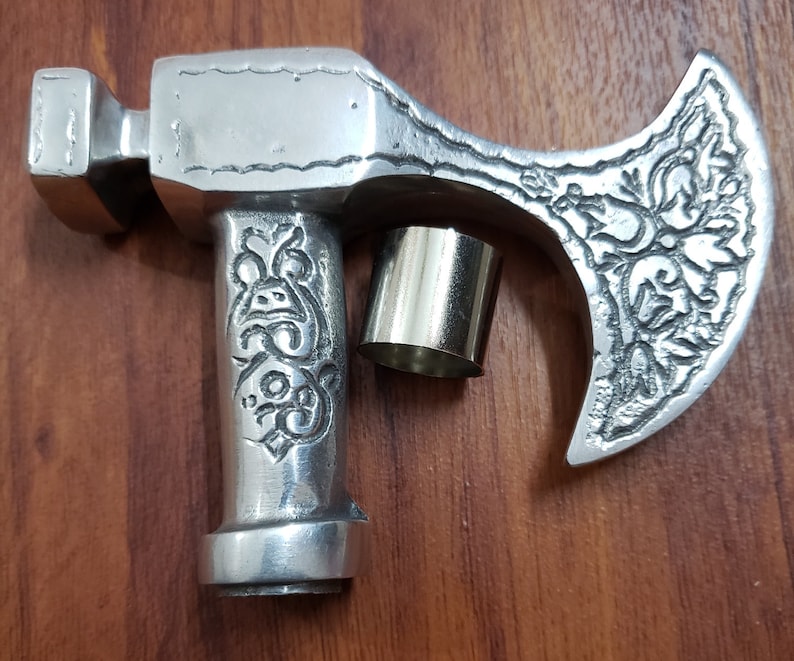 Puede incluir: Una cabeza de hacha de metal de color plateado con un dise&ntilde;o ornamentado. La cabeza del hacha est&aacute; unida a un mango de metal plateado con un dise&ntilde;o decorativo. La cabeza del hacha est&aacute; sobre una superficie de madera.