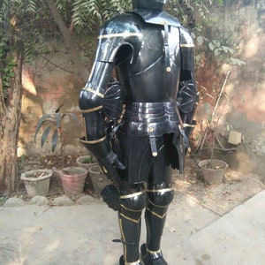 May include: A full suit of black armour with gold accents. The armour is made of metal and is designed to protect the wearer from weapons. The armour is standing on a wooden base.
