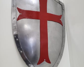 May include: A silver-colored shield with a red cross design. The shield has a riveted metal border and a curved shape. The red cross is centered on the shield.