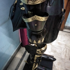 May include: Black and gold knight's armour with decorative details. The armour is made of metal and has a shiny finish. The armour is displayed on a black stand.