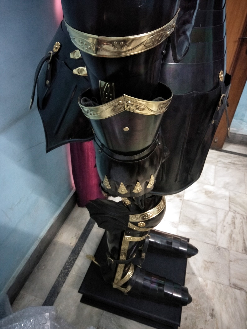 May include: Black and gold knight's armour with decorative details. The armour is made of metal and has a shiny finish. The armour is displayed on a black stand.