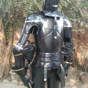 May include: A full suit of black armour with gold accents. The armour is made of metal and has a helmet, breastplate, gauntlets, and leg plates. The armour is standing on a dirt surface.