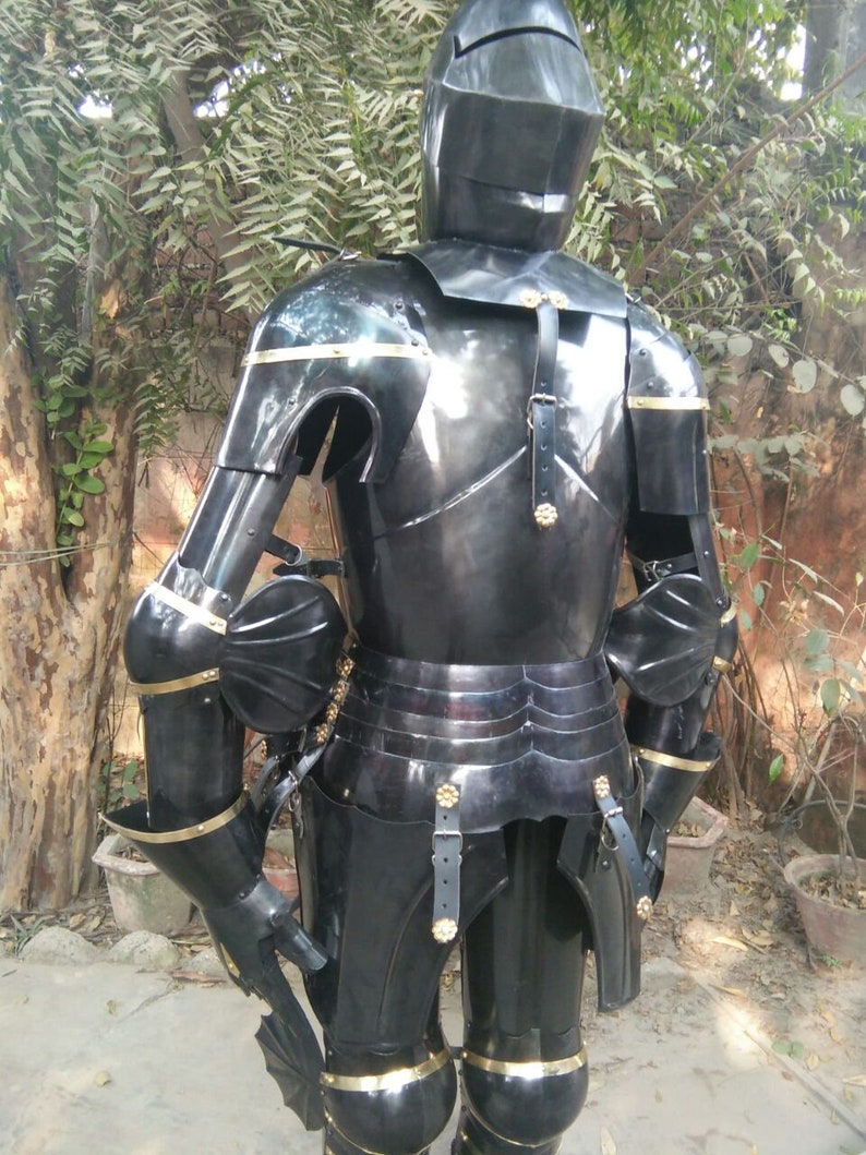 May include: A full suit of black armour with gold accents. The armour is made of metal and has a helmet, breastplate, gauntlets, and leg plates. The armour is standing on a dirt surface.