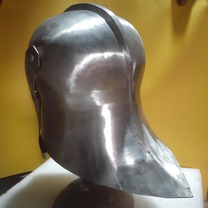 May include: A polished, silver-colored metal helmet with a rounded top and a curved visor. The helmet is mounted on a dark brown, rounded base. The helmet's surface reflects light, highlighting its smooth texture and design.