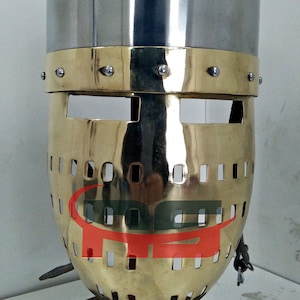 Puede incluir: Un casco medieval con una máscara facial de latón y una parte superior plateada. El casco tiene una banda decorativa alrededor de la parte superior y una cadena que cuelga de la parte inferior.