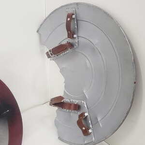 May include: A silver and red Captain America shield with a brown leather strap. The shield is slightly damaged, with a dent in the center.