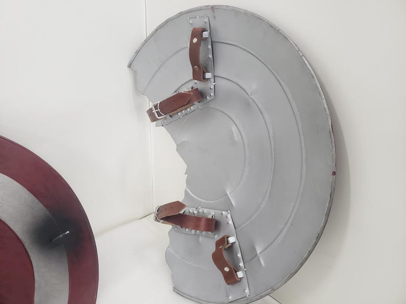 May include: A silver and red Captain America shield with a brown leather strap. The shield is slightly damaged, with a dent in the center.