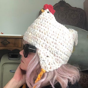 May include: A white crocheted chicken hat with a red comb and yellow yarn braids. The hat is being worn by a person with pink hair.
