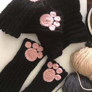 Könnte beinhalten: Ein Paar schwarze, fingerlose Strickhandschuhe mit rosa Pfotenabdrücken. Die Handschuhe sind aus weichem Garn gefertigt und perfekt, um Ihre Hände im Winter warm zu halten. Die Garnbälle sind ebenfalls im Bild enthalten.
