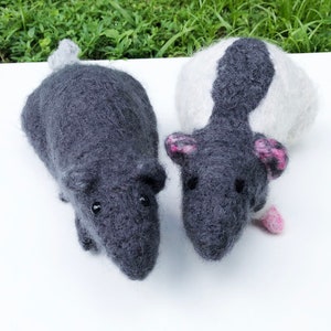 Puede incluir: Dos figuritas de rata de fieltro de aguja. Una rata es gris con una cola gris. La otra rata es blanca con una mancha negra en la espalda y orejas rosas.