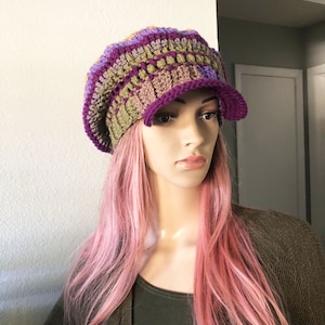 May include: A crocheted hat with a wide brim in shades of purple, green, and brown. The hat is worn by a mannequin with pink hair.