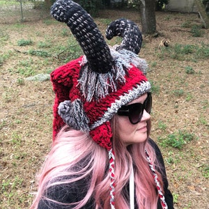 May include: A red crocheted hat with black and gray horns and a long red and white braided cord. The hat has a fluffy gray trim around the brim.