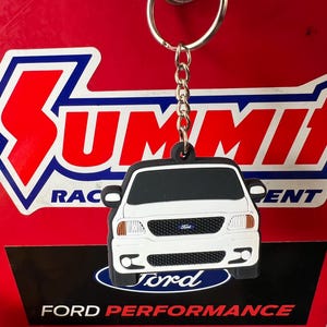 May include: White Ford F-150 truck keychain with a black background and the Ford logo. The keychain is attached to a silver key ring.