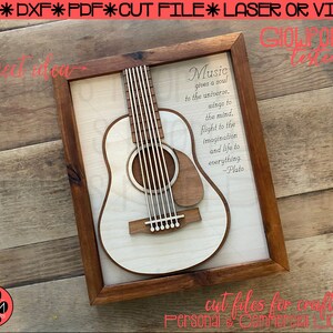 May include: A wooden frame with a white background and a cutout of an acoustic guitar. The quote "Music gives a soul to the universe. Wings to the mind, flight to the imagination and life to everything. -Plato" is printed on the background. The frame is on a wooden surface.
