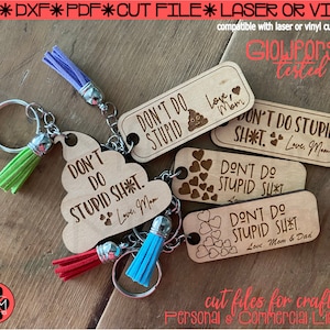 May include: Five wooden keychains with different designs and sayings. The keychains are laser cut and engraved with the phrases "Don't do stupid sh*t", "Love, Mom", "Love, Mom & Dad", and "Love, Mom". The keychains are on a wooden surface with a green, blue, and red tassel attached to each keychain.