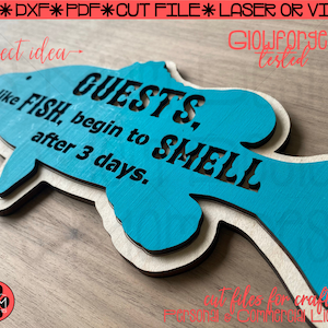 May include: A teal blue fish-shaped wooden sign with the text "Guests, Smell like Fish, begin to after 3 days." The sign is a project idea for crafters and is tested on a GlowForge.