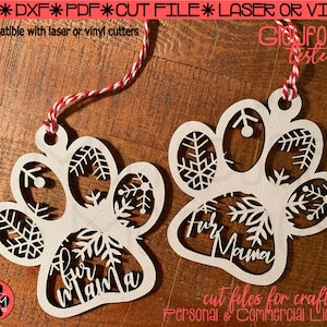 May include: Two white paw print ornaments with snowflake designs and the text "Fur Mama" cut out. The ornaments are hanging from red and white twine on a wooden surface. The text "cut files for crafters" and "Personal & Commercial License" is also visible.