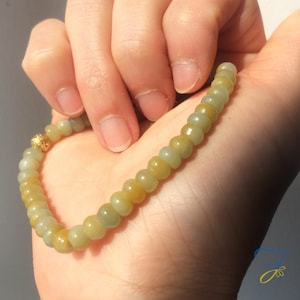May include: A beaded bracelet featuring small, round, light green and yellow beads. The bracelet has a gold-colored clasp. The beads are strung together to form a flexible strand, suitable for wearing around the wrist.
