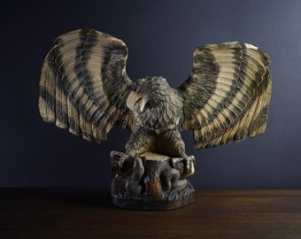 May include: A wooden carving of an eagle with its wings spread wide. The eagle is perched on a tree stump with its talons gripping the wood. The carving is painted in shades of brown and black.