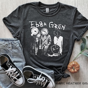 以下が含まれることがあります： ダークヘザーグレーのTシャツに、白い時計の頭をした4人の人物のグラフィックがプリントされています。人物の上に「Ebb & Grow」という文字がプリントされています。