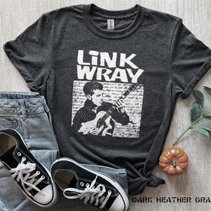 Puede incluir: Camiseta gris oscuro jaspeada con una imagen en blanco y negro de Link Wray tocando la guitarra. El texto "Link Wray" está impreso en blanco en la camiseta.