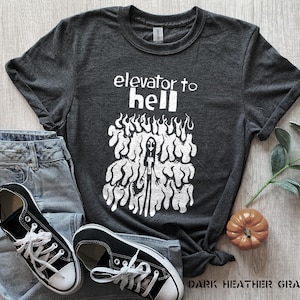 May include: Dark grey t-shirt featuring the text "elevator to hell" and a white graphic. The shirt is styled with blue jeans and black canvas trainers. A small orange pumpkin and greenery are also present.
