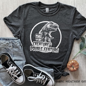 May include: Dark heather gray t-shirt with a white graphic of a dinosaur inside a circle, with the text "56 WLVI CREATURE DOUBLE FEATURE." The shirt is paired with blue jeans and black canvas sneakers.
