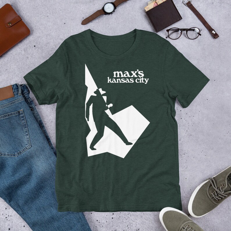May include: A dark green t-shirt with a white silhouette of a person in a hat walking. The text "max's kansas city" is printed in white on the shirt.
