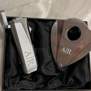 Puede incluir: Un juego de encendedor y cortador de cigarros plateado presentado en una caja negra con forro de sat&eacute;n negro. Tanto el encendedor como el cortador tienen las iniciales "A|R" grabadas. El encendedor es rectangular con tapa abatible y el cortador es de doble hoja.