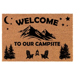 Op de afbeelding: Welkom op onze camping deurmat met een bruine kokosvezel achtergrond en zwarte tekst en afbeeldingen. De tekst luidt "Welkom op onze camping" met een bergketen, sterren, een halve maan, een kampvuur en twee stoelen.