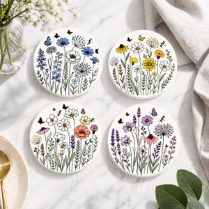 May include: Set of four round ceramic coasters with floral designs. Each coaster features a different arrangement of colorful flowers, butterflies, and dragonflies against a white background. The coasters are approximately 4 inches in diameter.