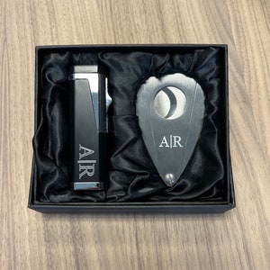 Puede incluir: Un juego de regalo negro que contiene un encendedor y un cortador de cigarros, ambos con las iniciales "A|R" grabadas. Los art&iacute;culos se presentan en una caja negra con un forro de sat&eacute;n. El encendedor tiene un acento plateado.