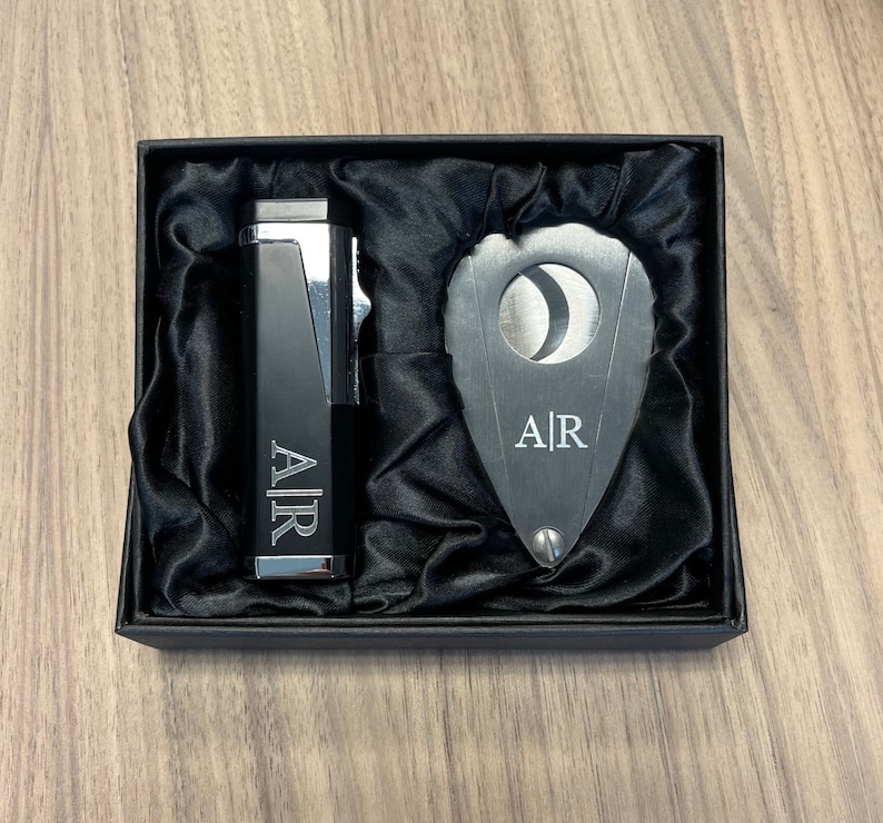 Puede incluir: Un juego de regalo negro que contiene un encendedor y un cortador de cigarros, ambos con las iniciales "A|R" grabadas. Los art&iacute;culos se presentan en una caja negra con un forro de sat&eacute;n. El encendedor tiene un acento plateado.