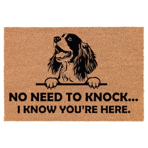 May include: A brown coir doormat with a black silhouette of a Springer Spaniel dog peeking over the top. The text "NO NEED TO KNOCK... I KNOW YOU'RE HERE." is printed in black below the dog.