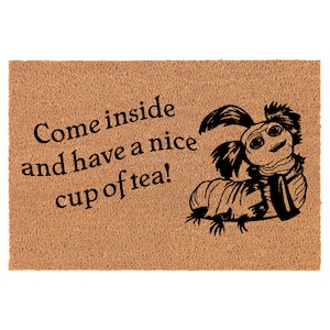 May include: A rectangular doormat with a light brown, textured surface. The mat features the text "Come inside and have a nice cup of tea!" in black, along with a black illustration of a caterpillar-like creature.