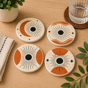 May include: Set of four round ceramic coasters with a cream background and orange and black celestial designs. Each coaster features a different sun, moon, and star pattern. The coasters are on a light wood surface.