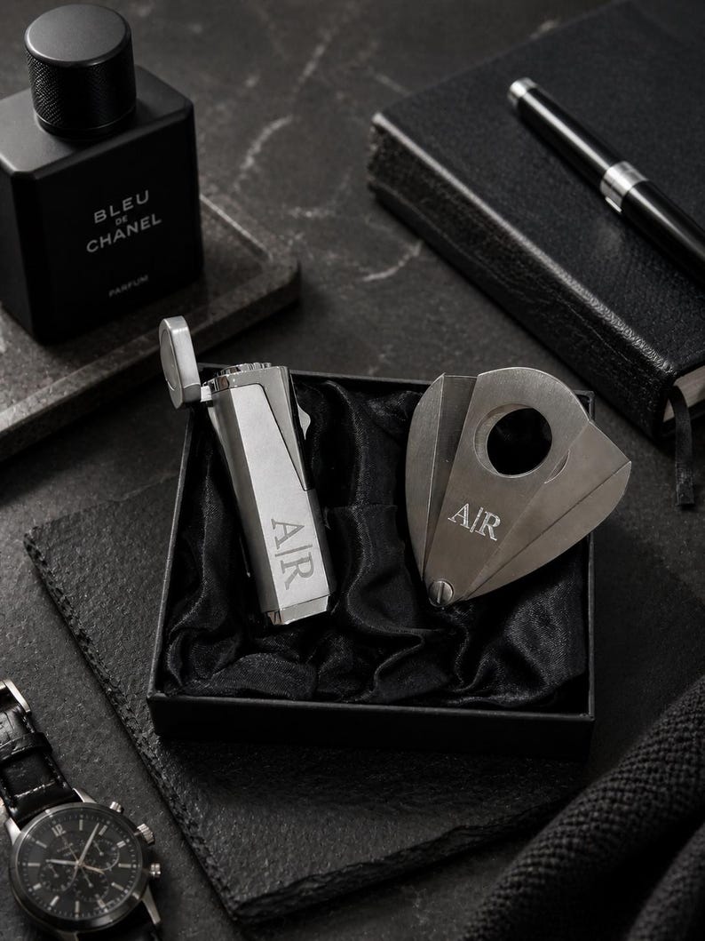 Puede incluir: Un lujoso juego de regalo con un cortador de cigarros y un encendedor plateados, ambos grabados con "A/R", presentados en una caja negra con forro de sat&eacute;n negro. Tambi&eacute;n se ven un reloj negro, un perfume "Bleu de Chanel", un bol&iacute;grafo y un cuaderno.
