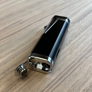 Puede incluir: Un encendedor negro y plateado con forma rectangular. El encendedor tiene una tapa con bisagras y una boquilla. El encendedor est&aacute; sobre una superficie de madera.