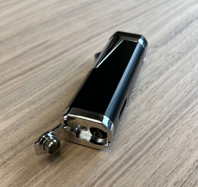 Puede incluir: Un encendedor negro y plateado con forma rectangular. El encendedor tiene una tapa con bisagras y una boquilla. El encendedor est&aacute; sobre una superficie de madera.