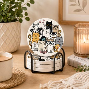 May include: A set of four ceramic coasters with a black metal holder. Each coaster features a different cartoon cat design in various colors. The coasters are stacked on a wooden surface, with a cup and candle in the background.