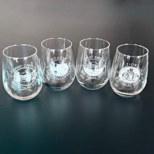May include: Four clear stemless wine glasses. Each glass features a different National Park design: Grand Canyon, Acadia, Great Smoky, and Zion. The park names and illustrations are printed in white.