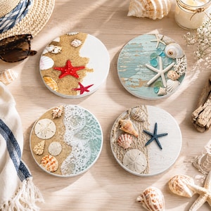 May include: Set of four round coasters with beach-themed designs. Each coaster features seashells, starfish, and sand, with variations including a distressed wood look and ocean wave details. The coasters are white and beige with red and blue accents.