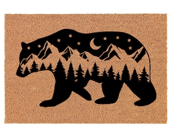 May include: A rectangular doormat with a brown background and a black bear silhouette. Inside the bear, a mountain range, trees, stars, and a crescent moon are visible. The mat is made of a natural fiber material.