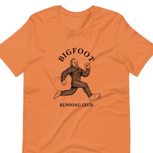 Pode incluir: Camiseta laranja com um gráfico preto de um Bigfoot correndo e o texto "BIGFOOT RUNNING CLUB".
