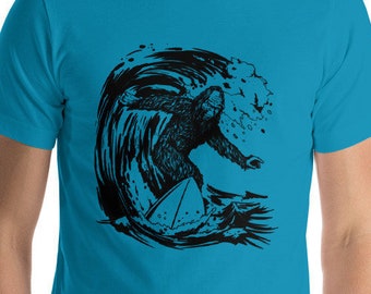 Puede incluir: Una camiseta azul turquesa con un gráfico negro de un Bigfoot surfeando en una ola. El gráfico incluye un pequeño pájaro volando sobre la ola.