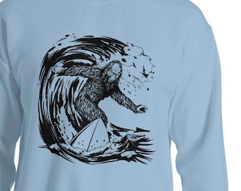 Puede incluir: Una sudadera de color azul claro con un gráfico en blanco y negro de un Bigfoot surfeando una ola.